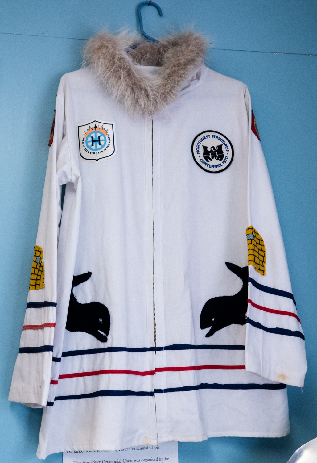A white parka like jacket hangs on a wall. The jacket has a fur collar, three stripes across the bottom and lower sleeves, two whales swimming toward each other, and symbolic patches.