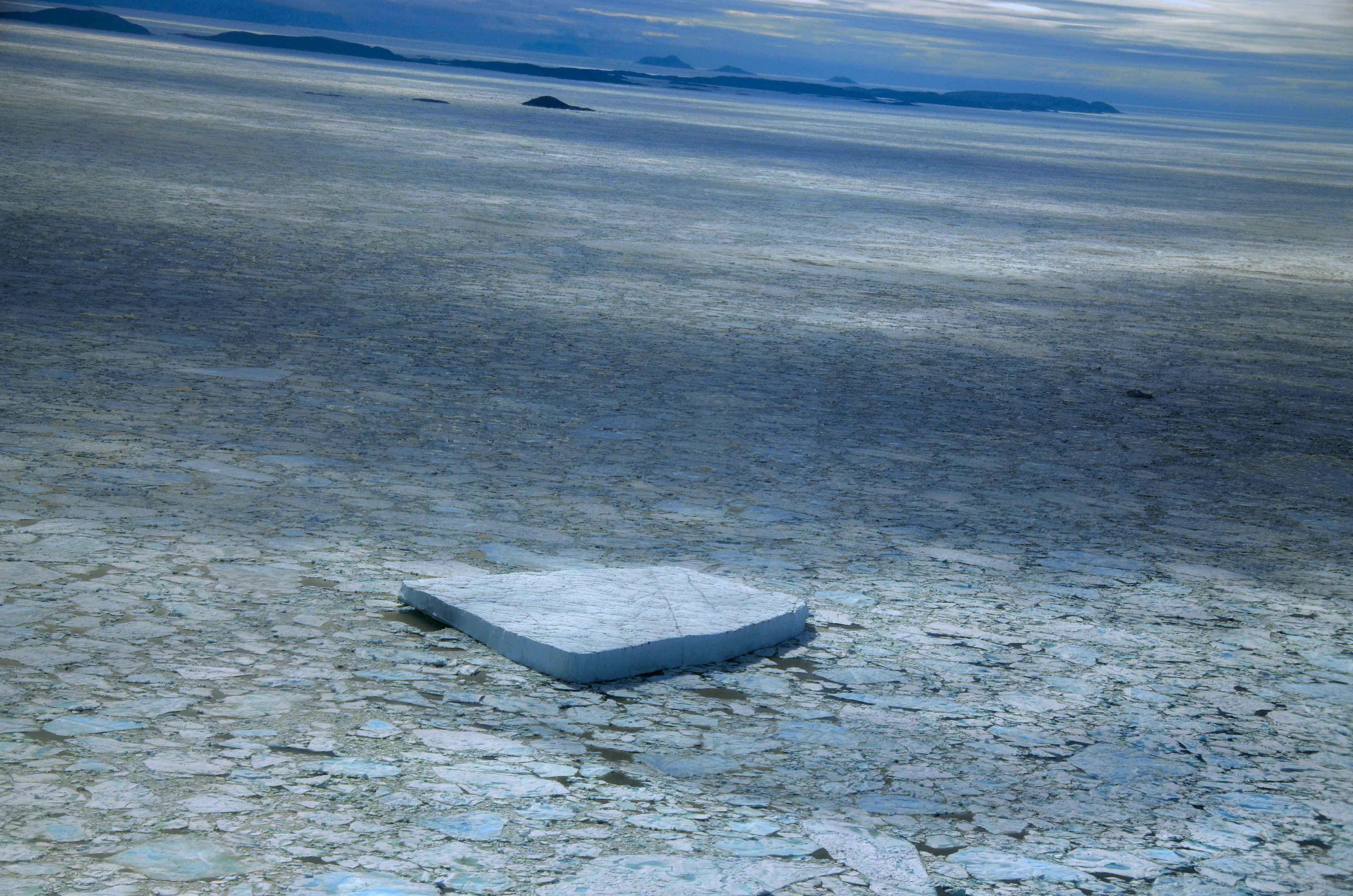 View from a low-flying plane of a sea covered in a mosaic of ice fragments, with a large, rectangular iceberg floating on top.