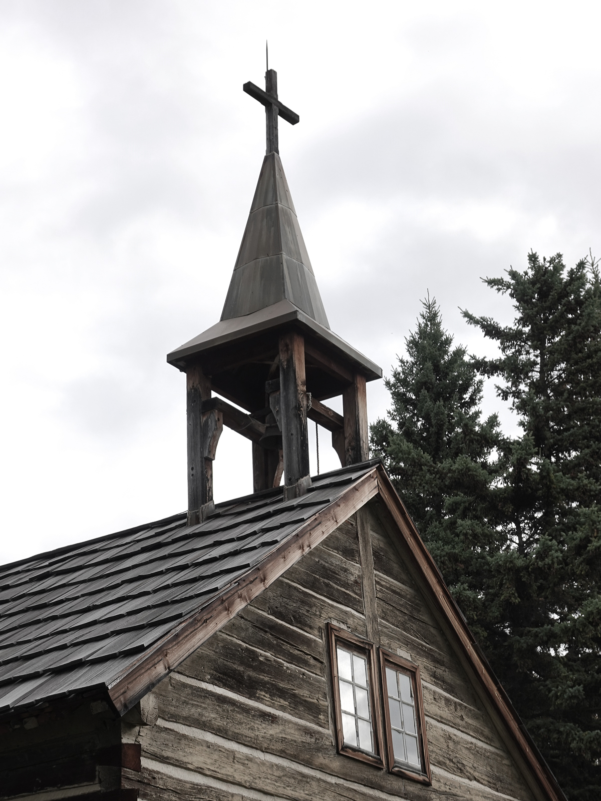 A cross and steeple rise above a wooden church building with fir trees as a backdrop.