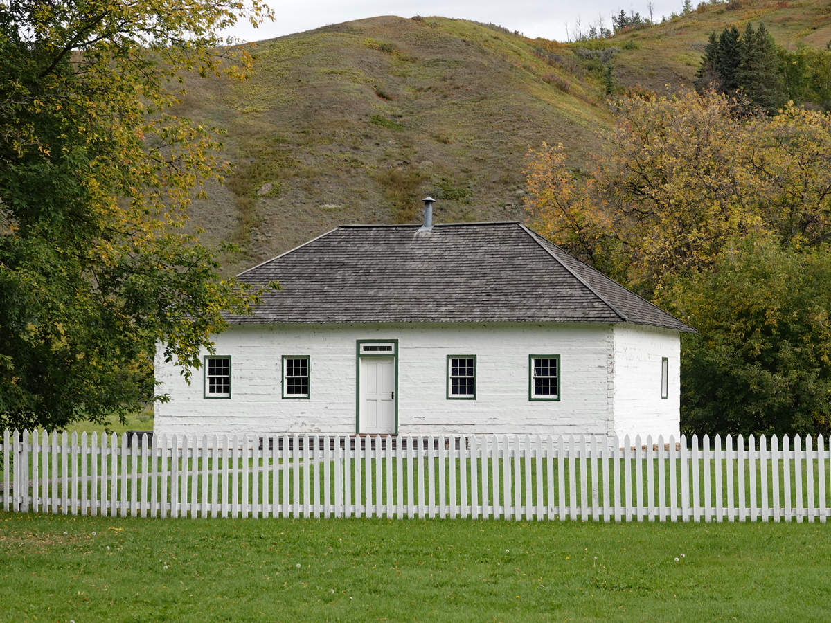 A small white building with tidy picket fence stands before a sandy hill.