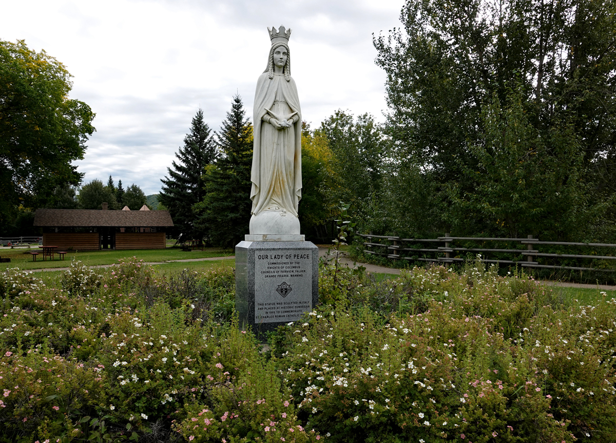 A white statue of the Virgin Mary wearing a crown rises above a pedestal with sign, surrounded by wild rose shrubs.