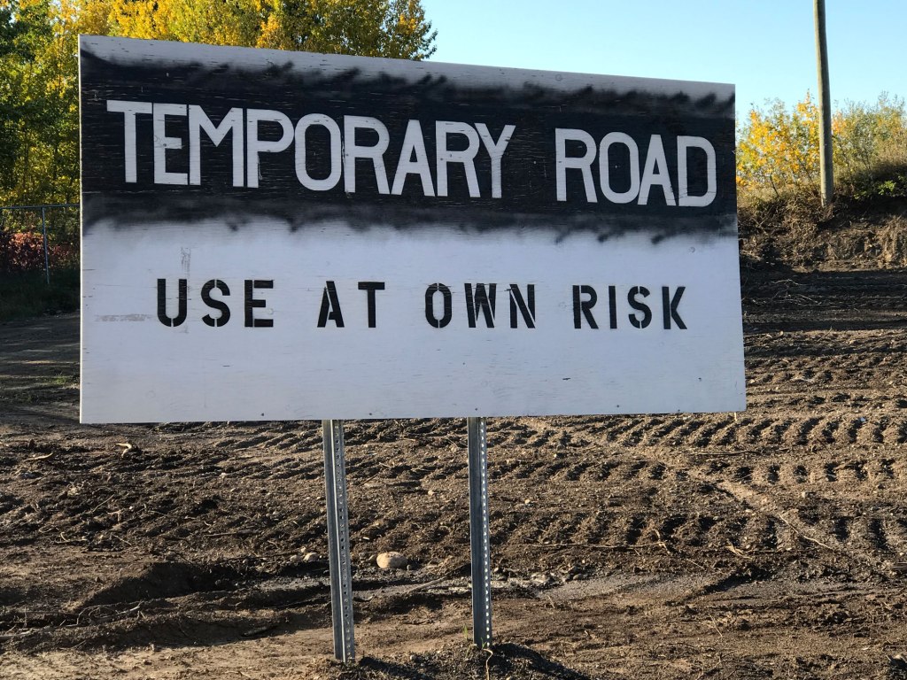 A black and white sign marks a graded brown dirt area. The sign says "Temporary Road: use at own risk".