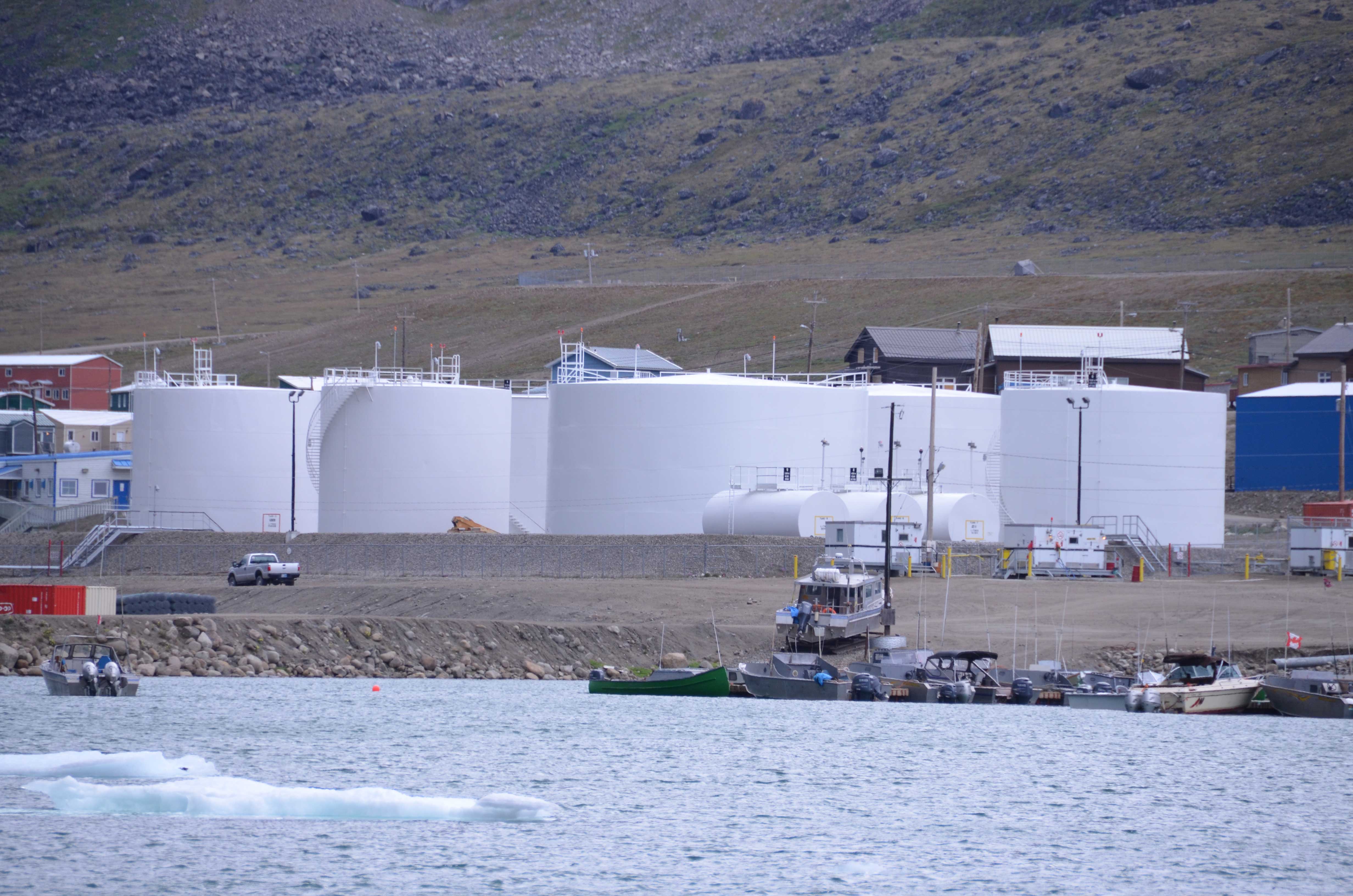Large white cylindrical tanks rise above the shore where boats are tied at a harbor.