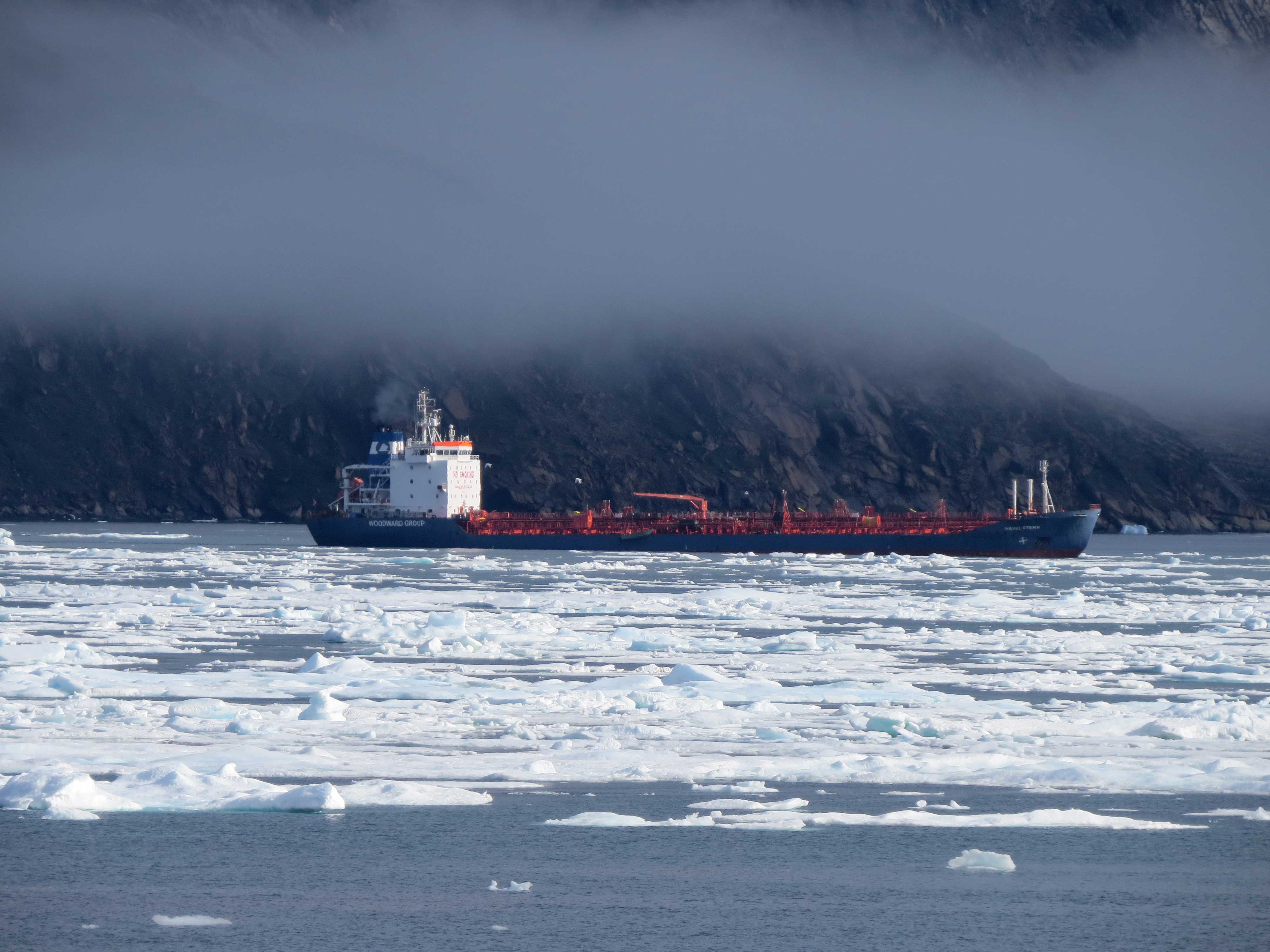 A long, dark blue tanker puffs smoke, with ice chunks across the waterway in front. A mountain disappears into fog behind it.