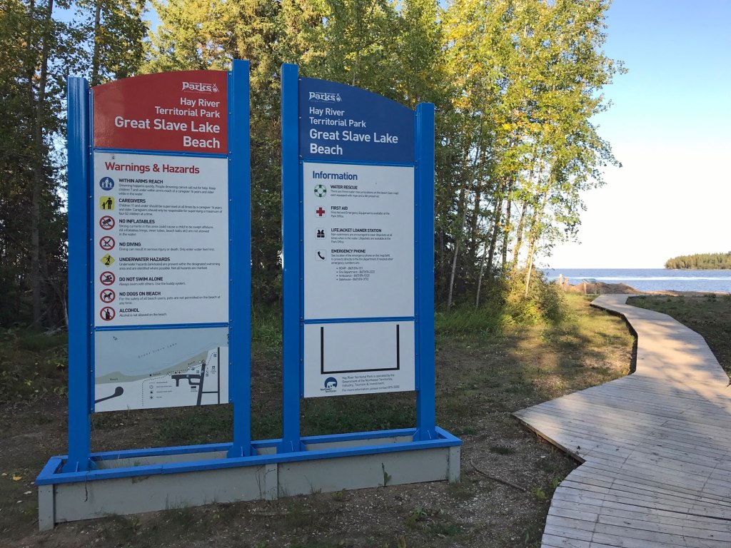 An elegant sign installation marks Great Slave Lake Beach at Hay River Territorial Park. A boardwalk runs past the blue sign mounts, leading to the water in the distance. 