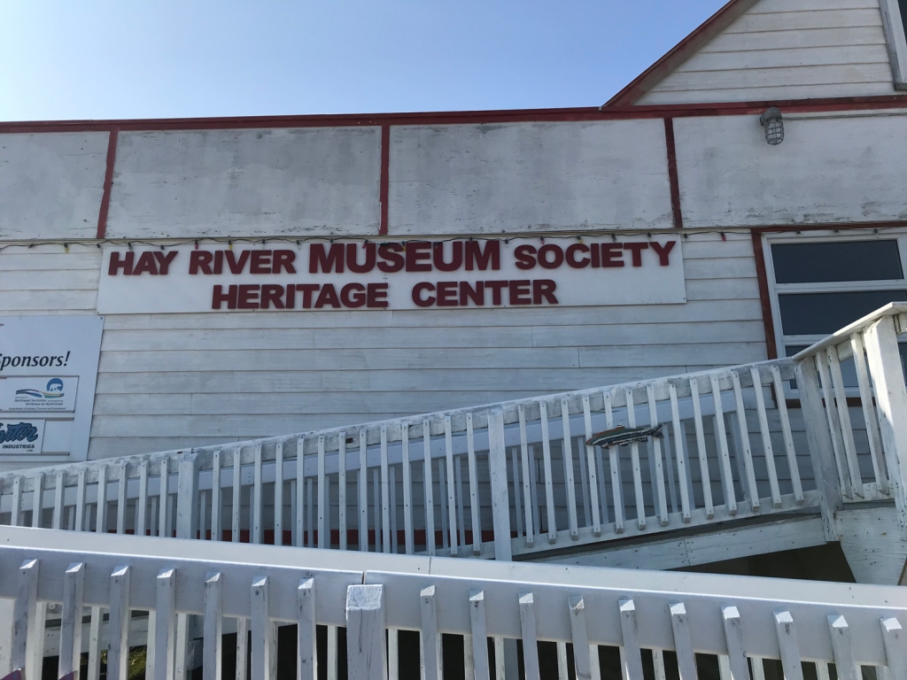 Entrance to the Hay River Heritage Center, showing a white painted wood ramp for accessibility, white siding, and red lettering. 
