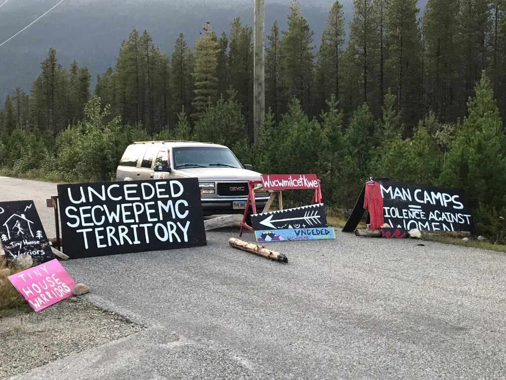 Image of a road blocked by an SUV and signs that say "unceded SECWEPEMC territory" and "man camps=violence against women". 