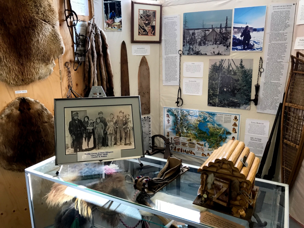 A booth wall and display case show gear and furs, along with photos, maps, and newspaper articles.