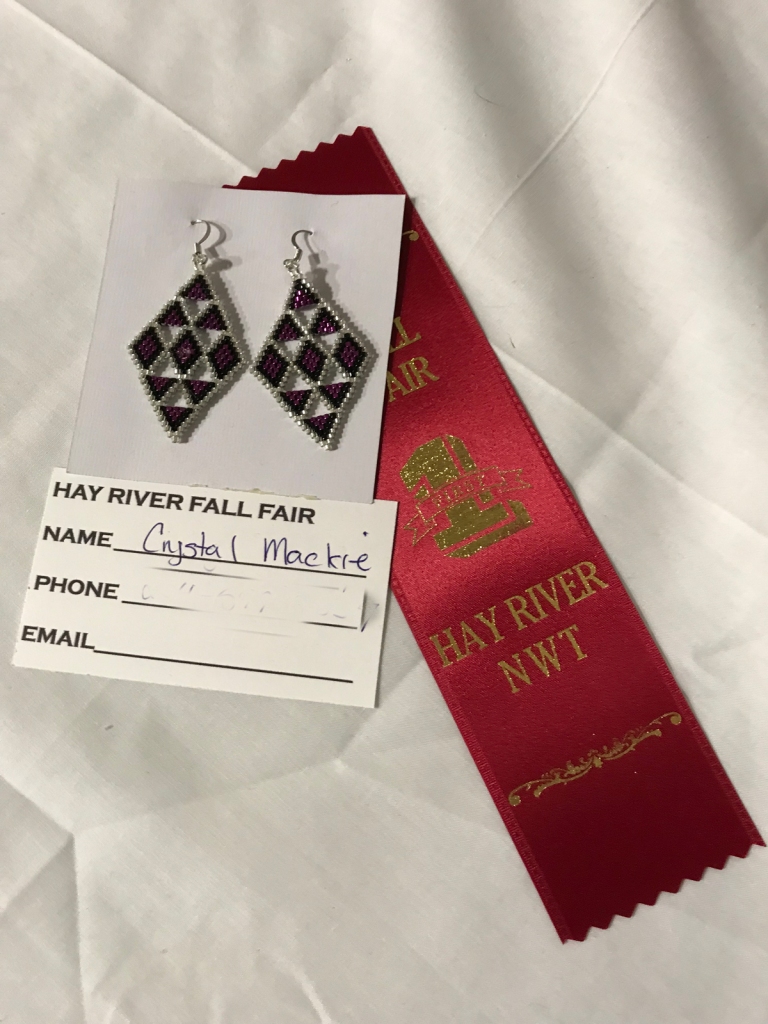Two beaded earrings are pinned to a card with an award ribbon lying underneath.
