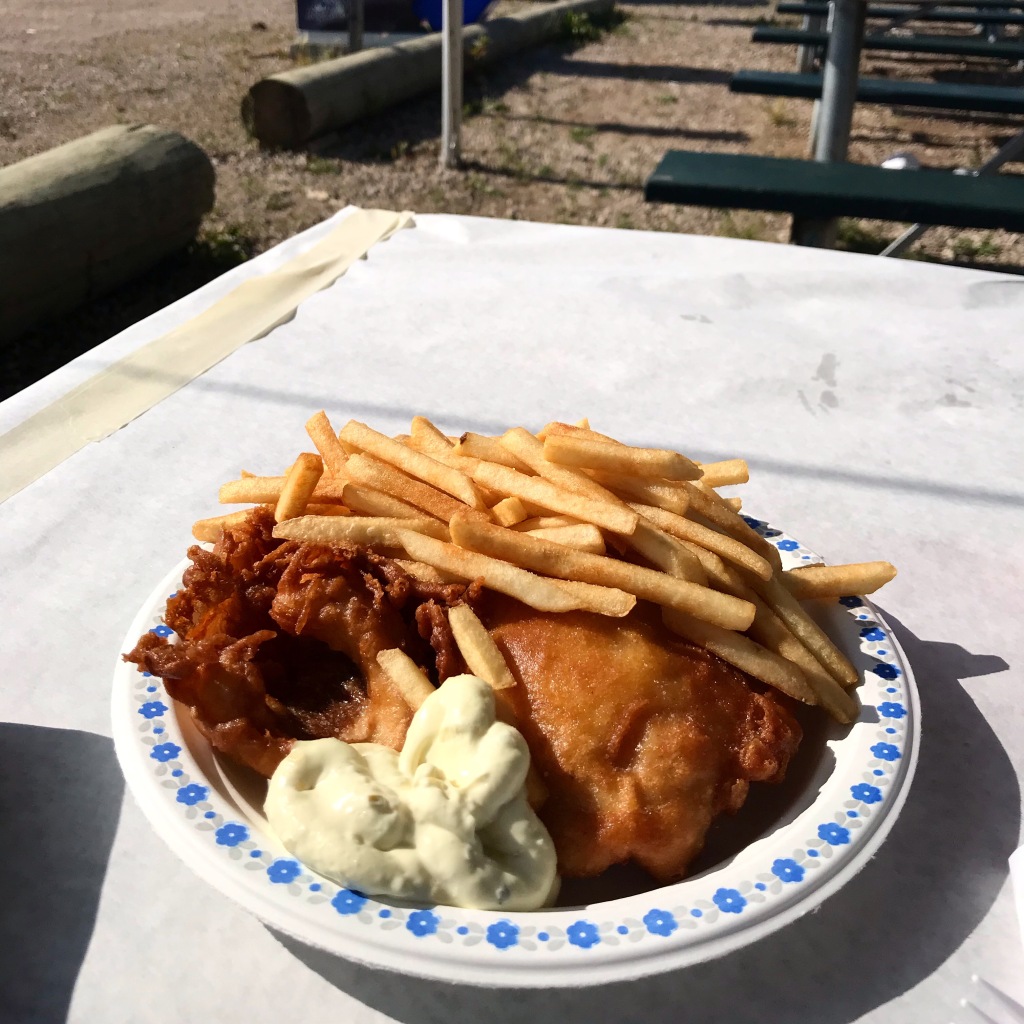 On top of a table with paper taped on top sits a full plate of beer-battered fish and chips with a healthy dollop of tartar sauce. 