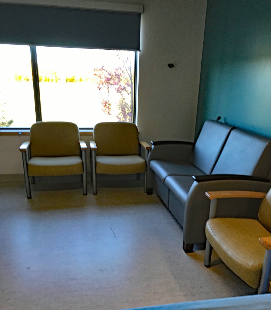 This photo shows yellow chairs and a grey couch in front of a window in a hospital exam room. 