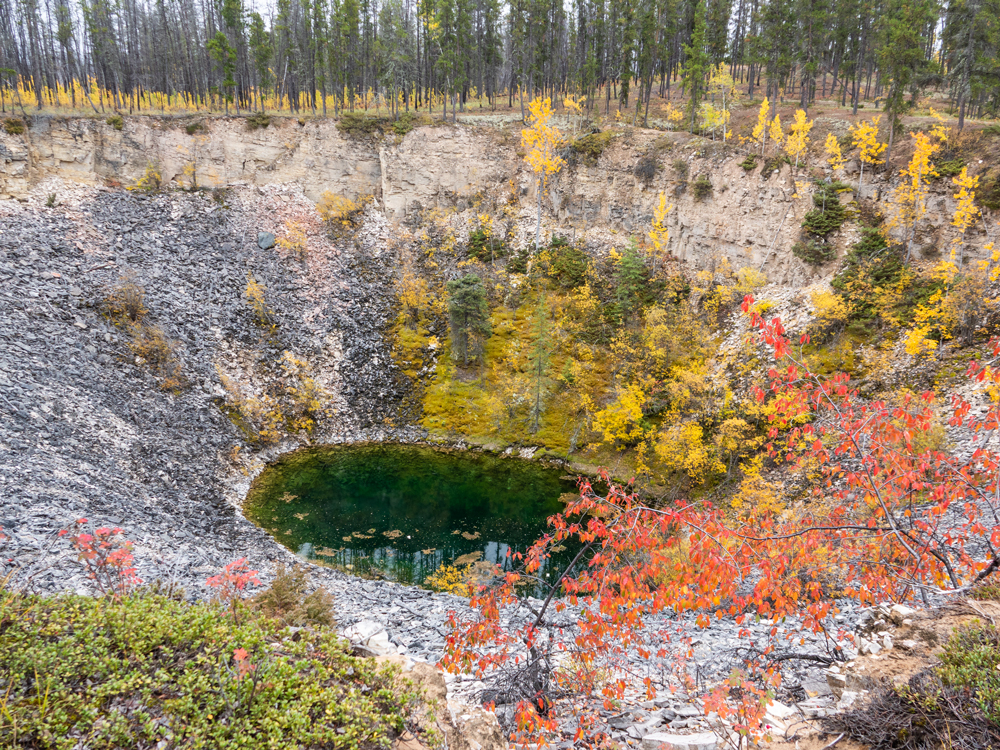 A circular water basin sits at the bottom of a deep funnel. The upper walls are tan with block lines running horizontally. Rocks and gravel cover the lower part, with some red, yellow, and green plants.